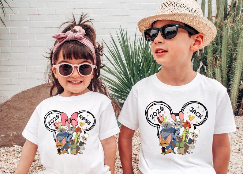 K&ouml;nnte beinhalten: Zwei Kinder tragen wei&szlig;e T-Shirts mit einer Cartoon-Grafik. Die Shirts zeigen ein Design mit dem Text "2026" und Charakterillustrationen. Ein Shirt hat den Namen "Rose", das andere "Jack". Der Junge tr&auml;gt eine Sonnenbrille und einen Strohhut.
