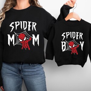 A woman wearing a black sweatshirt with the text "SPIDER MOM" and a smaller version of the same sweatshirt on a hanger.