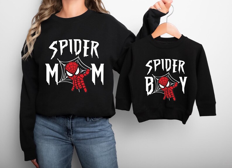 A woman wearing a black sweatshirt with the text "SPIDER MOM" and a smaller version of the same sweatshirt on a hanger.