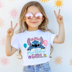 A young girl wearing a white t-shirt with a cartoon character and text, smiling and making a peace sign with her hands.