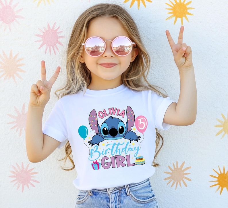 A young girl wearing a white t-shirt with a cartoon character and text, smiling and making a peace sign with her hands.
