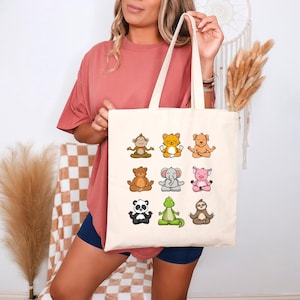 Może przedstawiać: Kremowa torba typu tote z dziewięcioma zwierzętami z kreskówek w pozycjach jogi. Zwierzęta to małpa, kot, pies, niedźwiedź, słoń, świnia, panda, aligator i leniwiec. Torba ma dwa kremowe uchwyty.