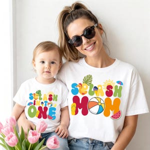 May include: White t-shirts with colorful text and graphics. One shirt says "Splash into One" with a beach theme, and the other says "Splash Mom". The shirts feature a beach ball, watermelon, and other summer elements.