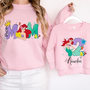 A woman wearing a pink sweatshirt with a mermaid design and a matching pink sweatshirt with a personalized name printed on it.