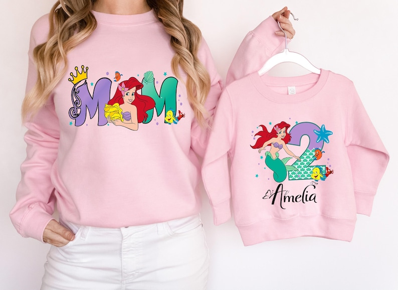 A woman wearing a pink sweatshirt with a mermaid design and a matching pink sweatshirt with a personalized name printed on it.