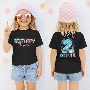 A young girl wearing a black t-shirt with a birthday message and a pink and white checkered hat, smiling and making a peace sign.