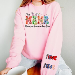A woman wearing a pink sweatshirt with a colorful design and text on it, sitting on a wooden stool and holding a white mug.