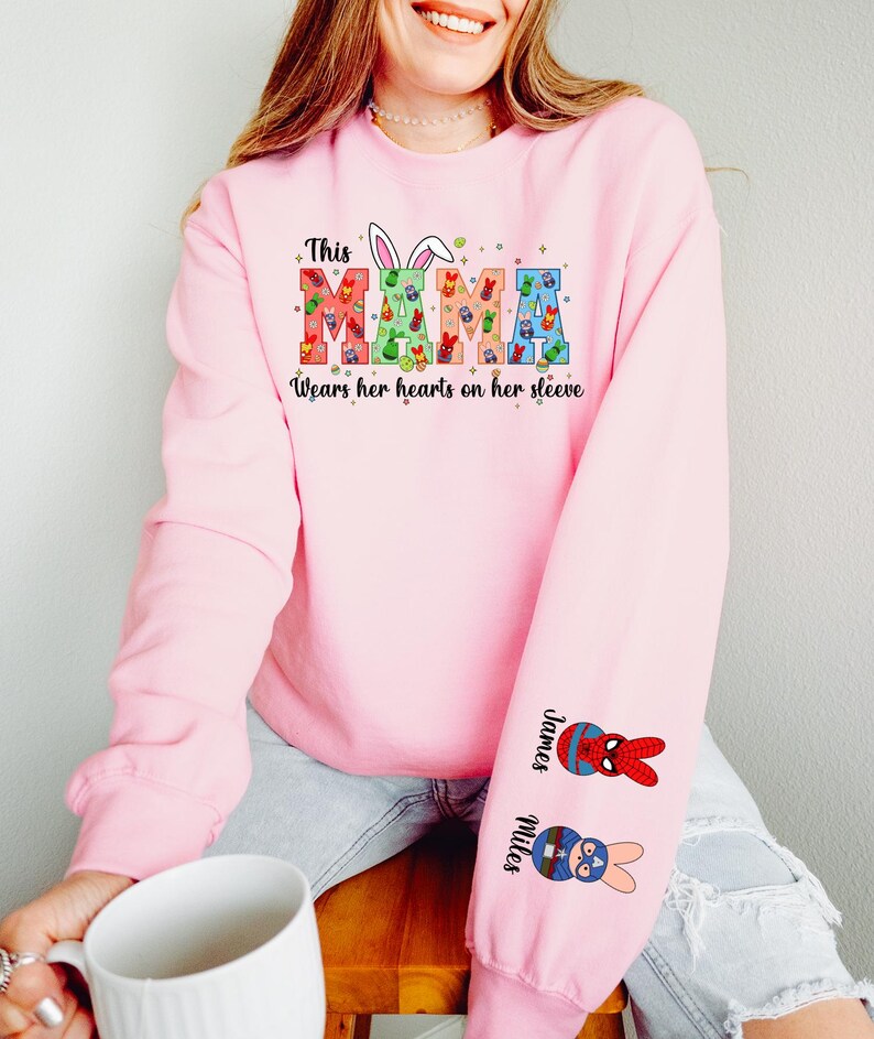 A woman wearing a pink sweatshirt with a colorful design and text on it, sitting on a wooden stool and holding a white mug.