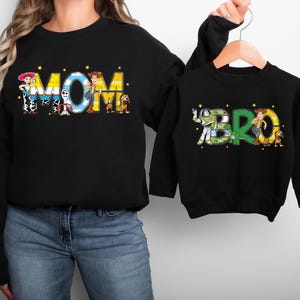 May include: Black sweatshirts with the words "MOM" and "BRO" in colorful, cartoon-style lettering. The "MOM" sweatshirt features characters from a popular animated film. The "BRO" sweatshirt is on a hanger.