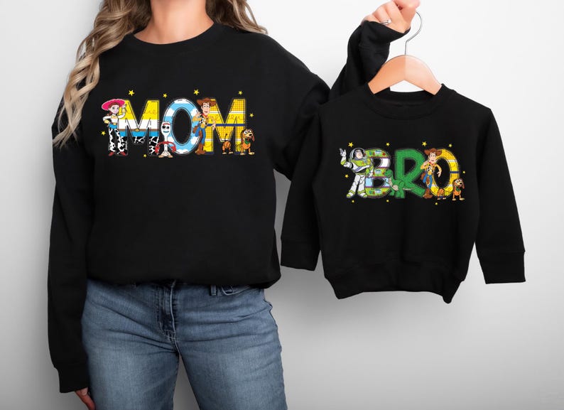 May include: Black sweatshirts with the words "MOM" and "BRO" in colorful, cartoon-style lettering. The "MOM" sweatshirt features characters from a popular animated film. The "BRO" sweatshirt is on a hanger.