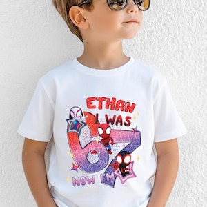 A young boy wearing a white t-shirt with a graphic design featuring Spider-Man characters and text.