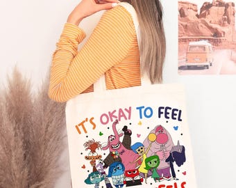 Könnte beinhalten: Eine cremefarbene Tragetasche mit einer bunten Grafik von Zeichentrickfiguren und dem Text "IT'S OKAY TO FEEL ALL THE FEELS" in Regenbogenfarben. Die Tasche wird von einer Person getragen, die ein gelb gestreiftes Oberteil trägt.