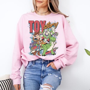 A woman wearing a pink sweatshirt with a colorful cartoon character design, including Woody from Toy Story, is standing next to a potted plant.