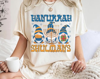 Könnte beinhalten: Cremefarbenes T-Shirt mit dem Text "Hanukkah with the Shulmans" in Blau und Orange. Das Design zeigt drei Zwerge mit blauen und orangenen Hüten und eine Menora. Die Zwerge halten Münzen, eine Menora und ein Geschenk.
