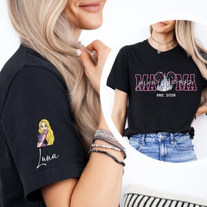 a woman wearing a black t-shirt with a cartoon character on the sleeve, and another woman wearing a black t-shirt with the text "MAMMA" and "EST. 2025" printed on it.