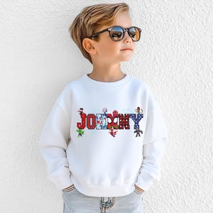 A young boy wearing a white sweatshirt with the word "JOMMY" printed on it, standing against a white wall.