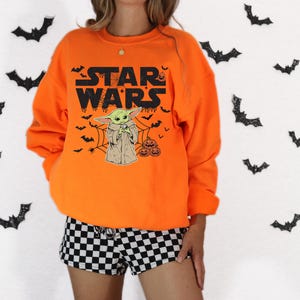 A person wearing an orange sweatshirt with a Star Wars character and bats printed on it, standing in front of a background with black bats.