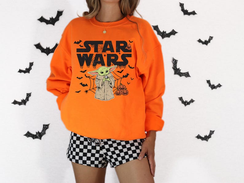 A person wearing an orange sweatshirt with a Star Wars character and bats printed on it, standing in front of a background with black bats.