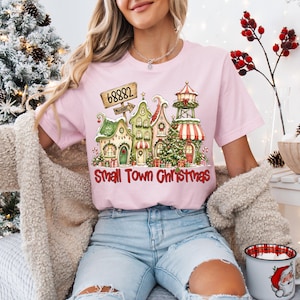 May include: A light pink short-sleeve t-shirt featuring a graphic print of a festive village scene with snow-covered houses, a Christmas tree, and a sign reading 68882. Below the illustration, the text Small Town Christmas is printed in red.