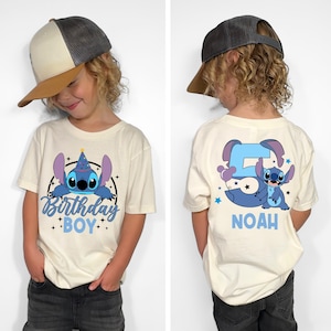 May include: Cream-colored t-shirt featuring a blue cartoon character with the text "Birthday Boy." The back of the shirt displays a large number 5 with the same character and the name "Noah" in blue. The child is wearing a gray and tan baseball cap.