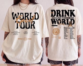 May include: An off-white t-shirt featuring black and gold graphic text. The front displays 'WORLD TOUR' with a list of countries. The back features 'DRINK AROUND THE WORLD' with a list of countries, cheers in various languages, and a globe icon.
