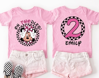 May include: Pink toddler t-shirts featuring Minnie Mouse. One shirt says "Oh Twoodles!" with Minnie Mouse making a peace sign. The other shirt has a pink number 2 and the name "Emily". Shown with white shorts and checkered shoes.