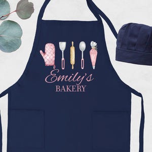 May include: Navy blue apron with two front pockets and a matching chef's hat. The apron features a watercolor design of baking tools and the text "Emily's Bakery" in pink script. A pink oven mitt, spatula, rolling pin, spoon, and pastry bag are depicted.