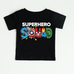 A black t-shirt with the text "SUPERHERO" printed on it, featuring various superhero logos and symbols such as Captain America, Spider-Man, Iron Man, and Hulk.