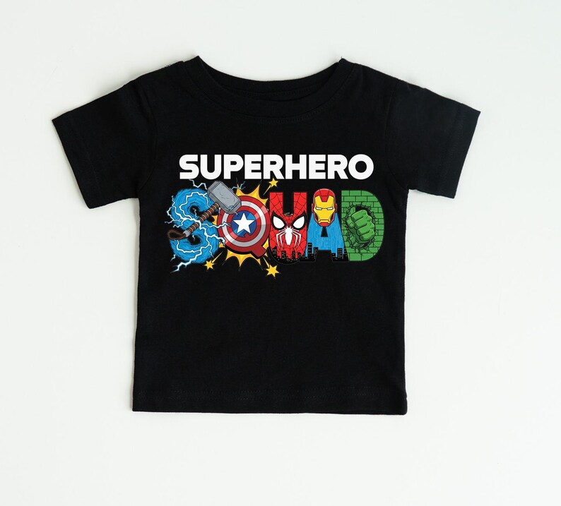 A black t-shirt with the text "SUPERHERO" printed on it, featuring various superhero logos and symbols such as Captain America, Spider-Man, Iron Man, and Hulk.