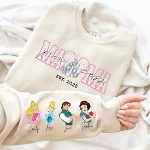 A person is holding a beige sweater with a Disney princess design and text printed on it.