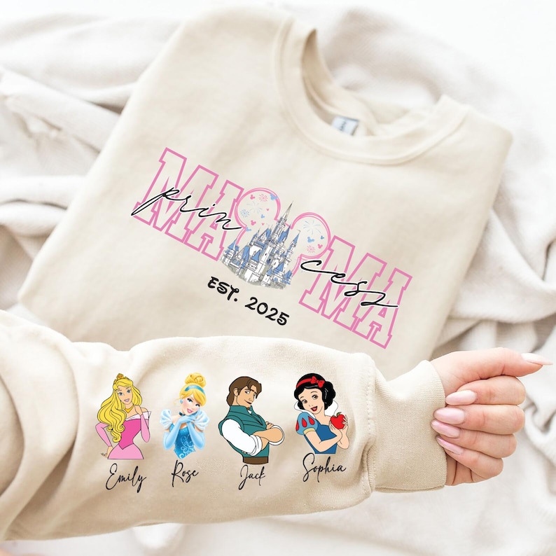 A person is holding a beige sweater with a Disney princess design and text printed on it.