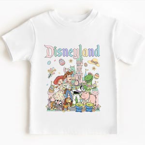Può includere: T-shirt bianca con grafica di Disneyland. Il design presenta la scritta "Disneyland" in colori pastello, insieme a personaggi dei cartoni animati come Woody, Jessie e Buzz Lightyear. La maglietta è per bambini e presenta personaggi di Toy Story.