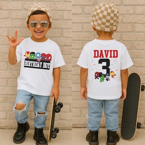 Op de afbeelding: Een wit T-shirt met een kleurrijke afbeelding van Marvel-superhelden en de tekst "BIRTHDAY BOY". De achterkant van het shirt heeft de naam "DAVID" en het nummer "3". Het kind draagt een blauwe jeans, zwarte laarzen en een beige-witte geruite muts.