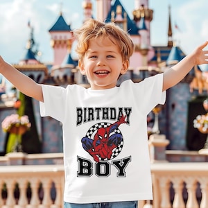 A young boy wearing a white t-shirt with a Spiderman graphic and the text "Birthday Boy" is smiling and raising his arms in front of a colorful, castle-like structure.