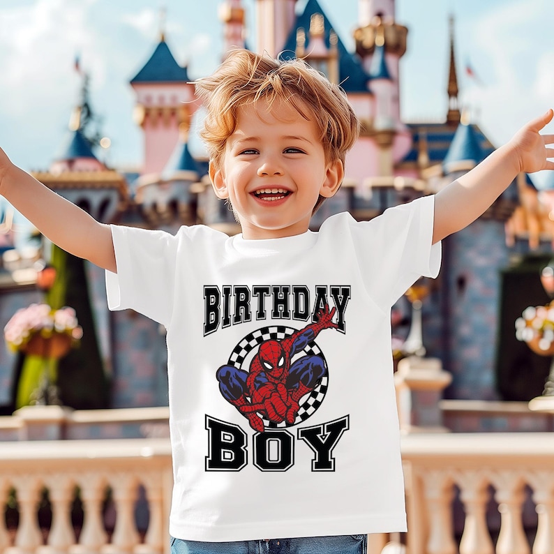 A young boy wearing a white t-shirt with a Spiderman graphic and the text "Birthday Boy" is smiling and raising his arms in front of a colorful, castle-like structure.