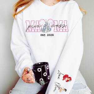 A woman wearing a white sweatshirt with a Disney-themed design and text, holding a black mug with a floral pattern.