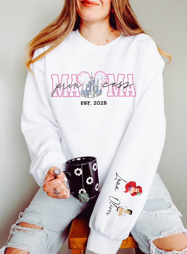 A woman wearing a white sweatshirt with a Disney-themed design and text, holding a black mug with a floral pattern.