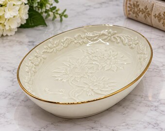 Könnte beinhalten: Eine ovale Keramikschale in cremeweißer Farbe mit einem erhabenen Blumenreliefmuster auf dem Innenboden und entlang des Randes. Der Rand der Schale ist mit einer dünnen, metallisch goldenen Verzierung versehen, die für ein edles Aussehen sorgt.