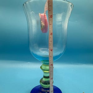 May include: A tall, clear glass goblet with a blue base and rim, a green stem, and a flared top. A pink measuring tape is inside the glass, showing a height of approximately 6 inches.