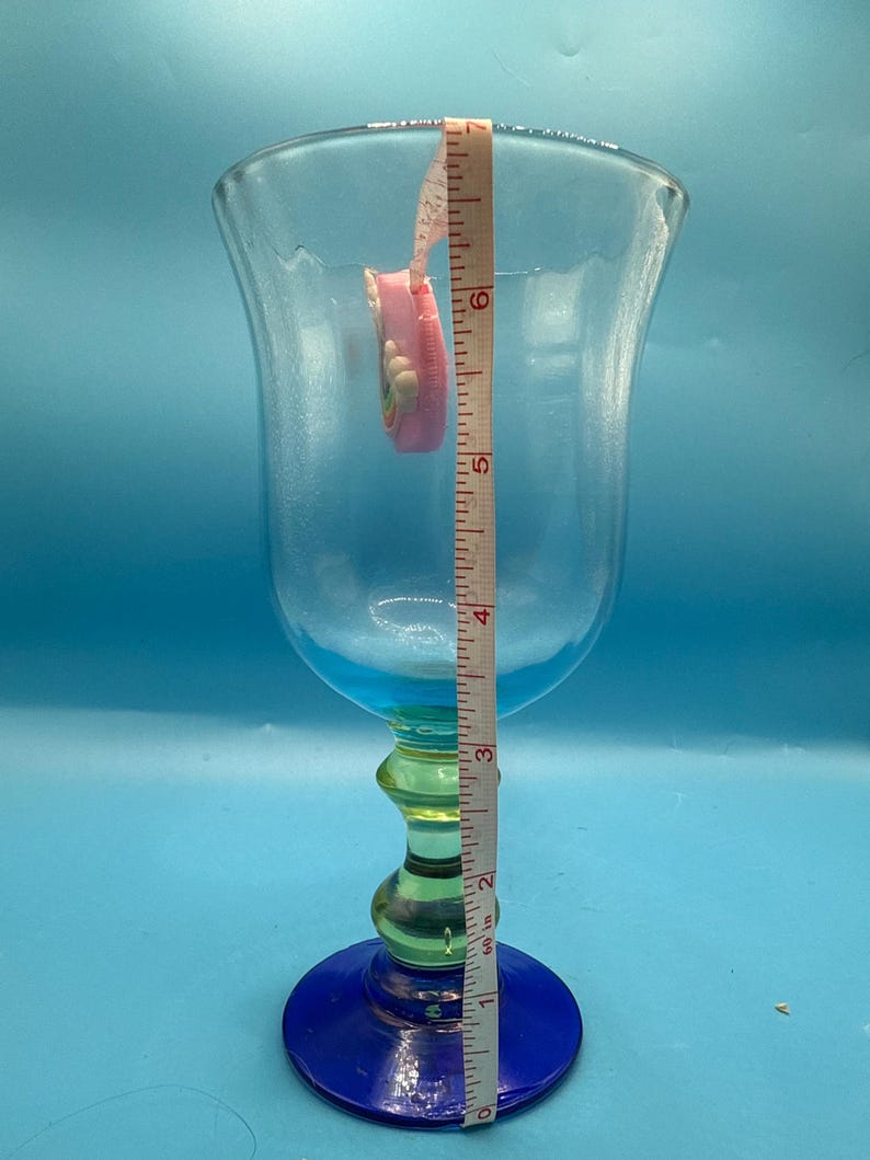 May include: A tall, clear glass goblet with a blue base and rim, a green stem, and a flared top. A pink measuring tape is inside the glass, showing a height of approximately 6 inches.