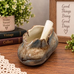 May include: A ceramic ashtray with a mottled brown and blue glaze, featuring an irregular, organic shape with a smooth cream-colored interior and a single integrated cigarette rest extending from the rim, showcasing a glossy, speckled finish.