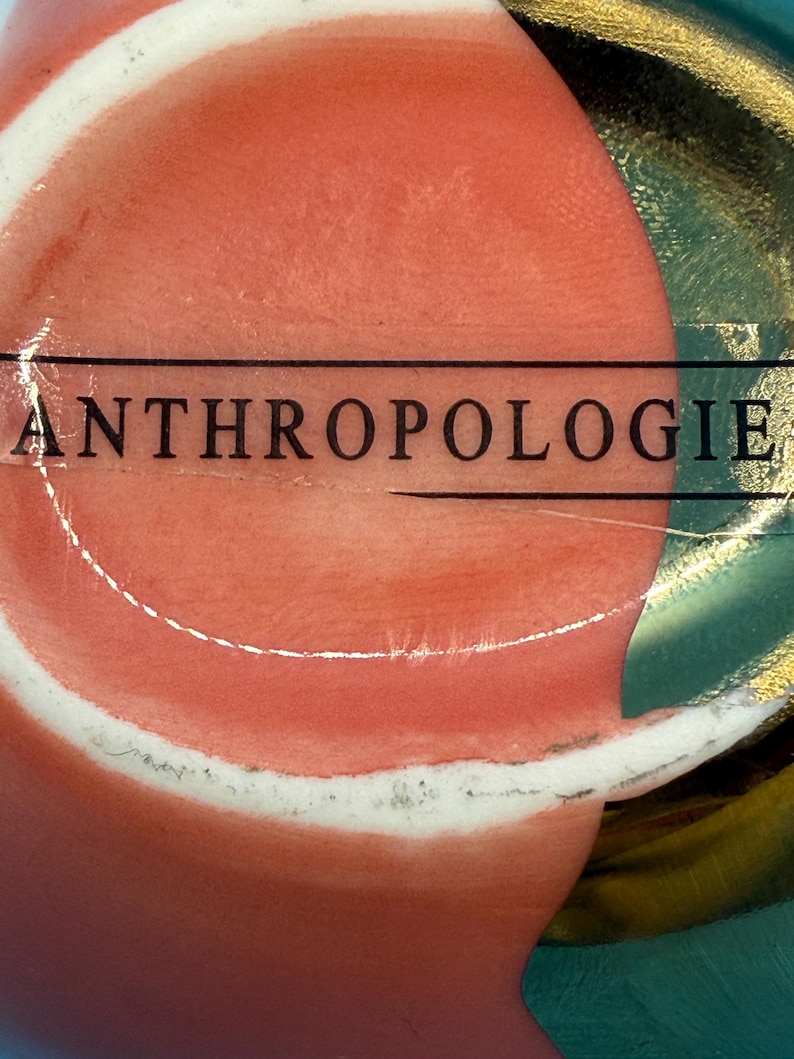 Puede incluir: Primer plano de un objeto decorativo con una superficie de color coral y un borde blanco. La palabra "ANTHROPOLOGIE" est&aacute; impresa en negro en el objeto. El fondo presenta detalles en verde azulado y dorado.