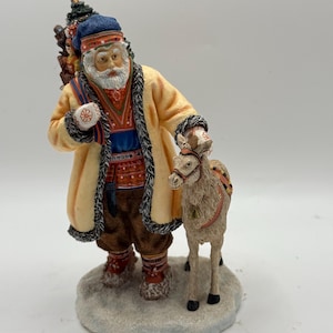 May include: A figurine of Santa Claus in a cream-colored coat with black trim, a blue hat, and a white beard, standing next to a small reindeer. Santa carries a pack on his back and holds a decorative ball. The figurine is on a snowy base.