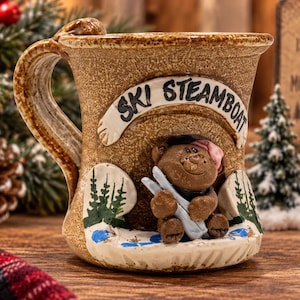 May include: A tan ceramic mug featuring a raised relief design of a brown bear wearing a pink hat and holding white skis. The text "SKI STEAMBOAT" is embossed in black letters on a curved banner above the bear, with painted green pine trees.