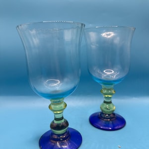 May include: Two clear glass goblets with blue rims and bases, and yellow and green stems. The goblets have a tulip shape and are set against a light blue background. The glassware is likely for serving beverages.