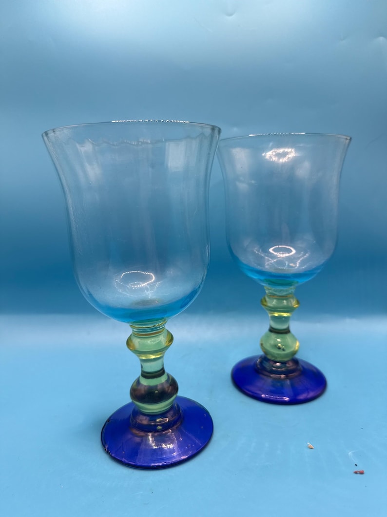 May include: Two clear glass goblets with blue rims and bases, and yellow and green stems. The goblets have a tulip shape and are set against a light blue background. The glassware is likely for serving beverages.