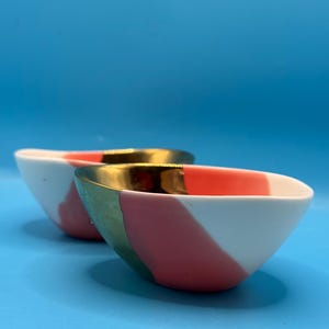 Puede incluir: Dos cuencos de cer&aacute;mica con una combinaci&oacute;n de colores blanco, rosa y verde. Los cuencos tienen un borde dorado y un dise&ntilde;o de bloques de color diagonal. Los cuencos est&aacute;n sobre un fondo azul.