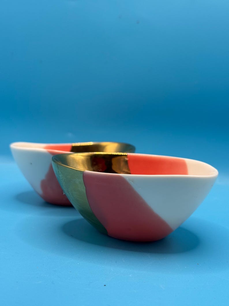 Puede incluir: Dos cuencos de cer&aacute;mica con una combinaci&oacute;n de colores blanco, rosa y verde. Los cuencos tienen un borde dorado y un dise&ntilde;o de bloques de color diagonal. Los cuencos est&aacute;n sobre un fondo azul.