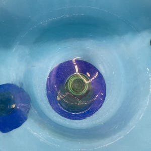 May include: A close-up view of a light blue ceramic bowl with a dark blue and green center. The bowl has a circular design with concentric rings of color. The interior of the bowl is smooth and glossy.