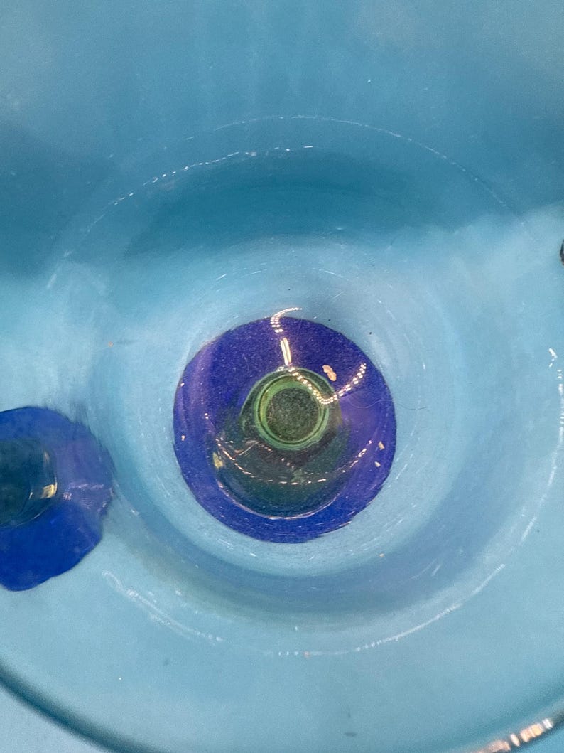 May include: A close-up view of a light blue ceramic bowl with a dark blue and green center. The bowl has a circular design with concentric rings of color. The interior of the bowl is smooth and glossy.